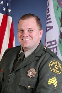 Image ID: A picture of Tyson Pogue, Madera County Sheriff-Coroner, a smiling man posing for a picture in uniform. There is an American and California Republic flag in the background. End ID.