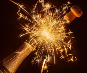 Image of a bottle of champagne being popped with a sparkler in the background. 