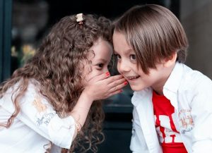 Image of a young girl whispering into the ear of a young boy. 