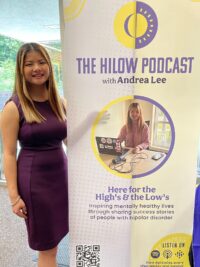 Andrea Lee standing next to a sign for the HiLow Podcast. 