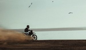 Image of a person riding a motorcycle in the sand with water in the background.