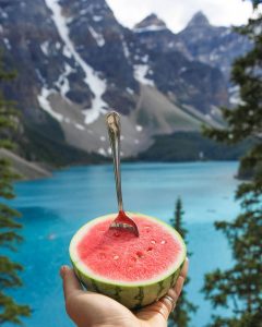 Image of half a watermelon with a spoon in it in front of a mountain lake.