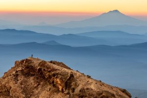 Image of a person standing on a mountain at sunset with several mountain ranges off in the distance.