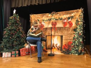 Image of an older gentleman reading a Christmas story in front of a fake fireplace and a christmas tree with presents