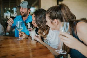 Photo of people drinking wine with a dog at the table mleming. aka sticking is tongue out on his nose