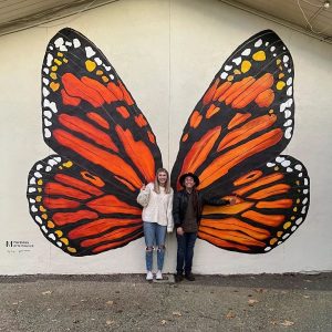 Image of two people standing in front of a butterfly mural. 