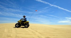 Image of someone riding and dune buggy along a sandy beach with blue sky in the background.