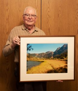 Gene Bryan is holding his “Golden Meadow” photograph that perfectly captures the beauty of the June Lake area in the fall, where the golden meadow, clear water, and towering mountains create a stunning and awe-inspiring landscape.